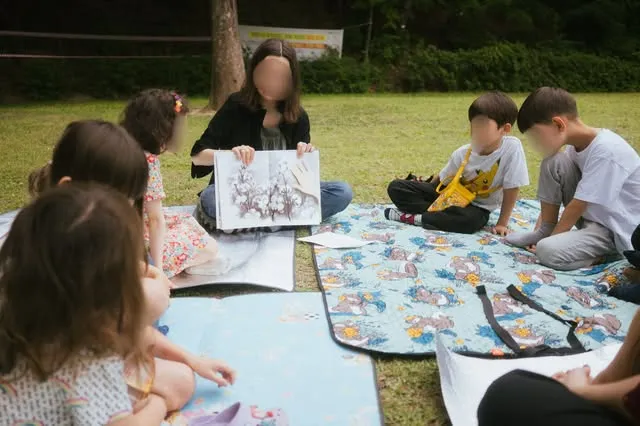Des enfants dessinent sur une nappe dans l'herbe, avec une artiste.